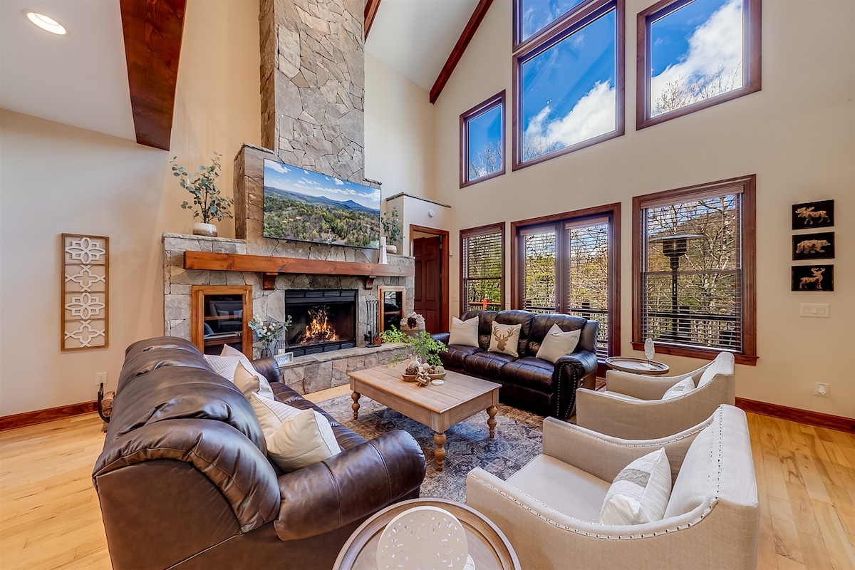 A spacious living area features a stone fireplace that serves as the focal point, with large windows allowing natural light to illuminate the room. Comfortable leather sofas and light-colored armchairs are arranged around a central coffee table, offering a cozy atmosphere complemented by decorative elements.