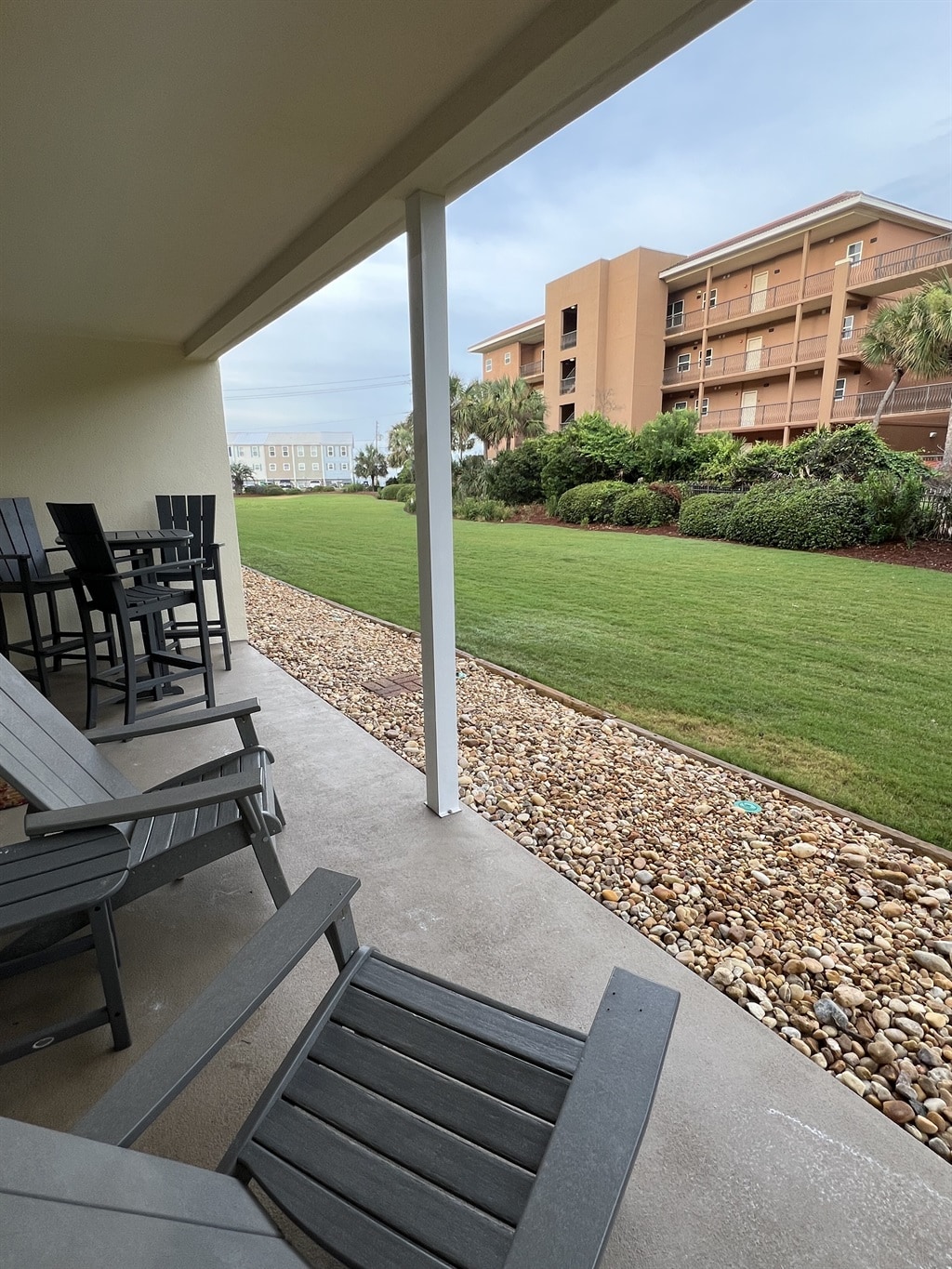 A relaxed outdoor seating area features dark wooden chairs and a table, positioned under a covered porch. Lush green grass stretches out beyond, bordered by neatly arranged stones and colorful landscaping. The building in the background adds depth to the serene environment.