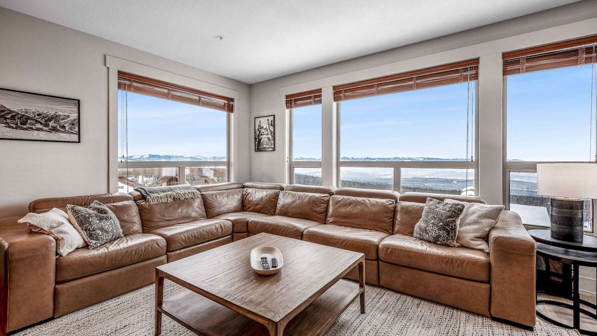 A spacious living room is highlighted by large windows offering panoramic views of the outdoors. A sectional leather sofa surrounds a wooden coffee table. Natural light fills the room, while warm tones in the decor create a cozy atmosphere.