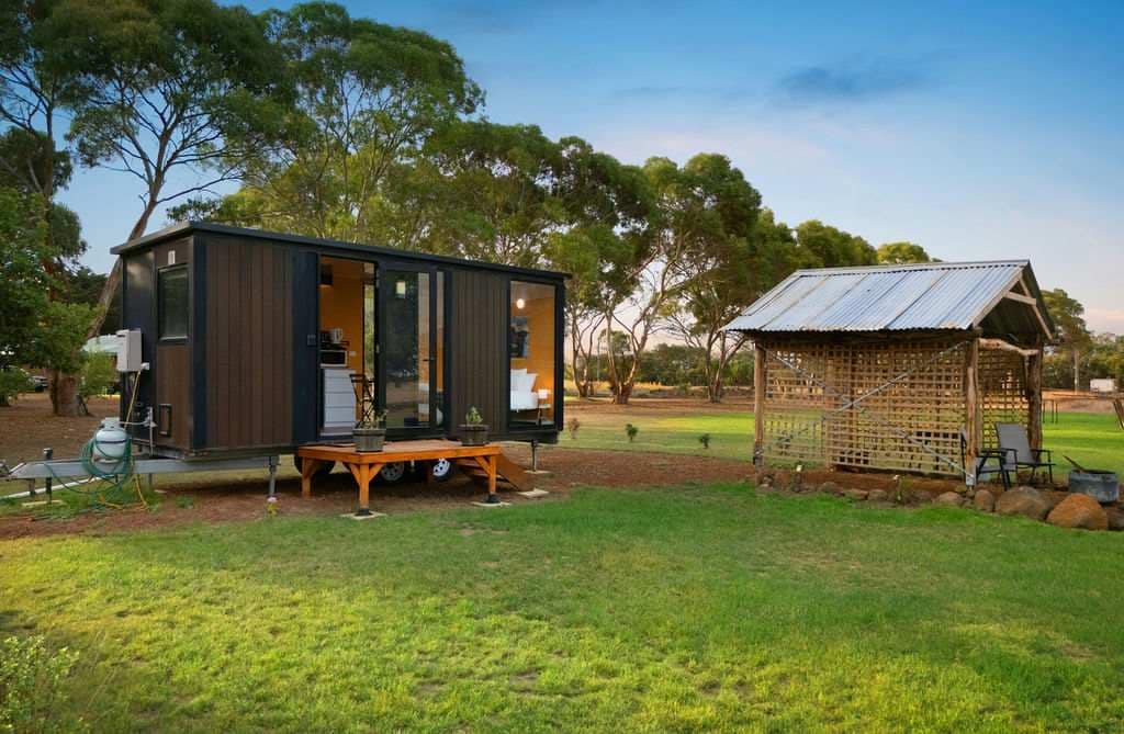 A tiny house is situated on grassy land, showing a welcoming outdoor patio with a wooden table. Nearby, a rustic shelter is visible, surrounded by trees and open fields, providing a serene rural atmosphere.