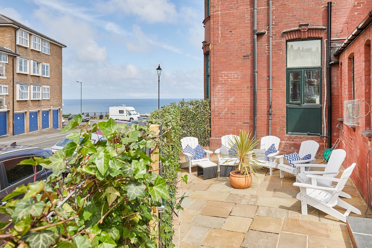 An outdoor terrace is presented with several white adirondack chairs arranged around a small coffee table. Lush greenery frames the space, offering a glimpse of the sea view in the background, while a blue sky with scattered clouds enhances the inviting atmosphere.