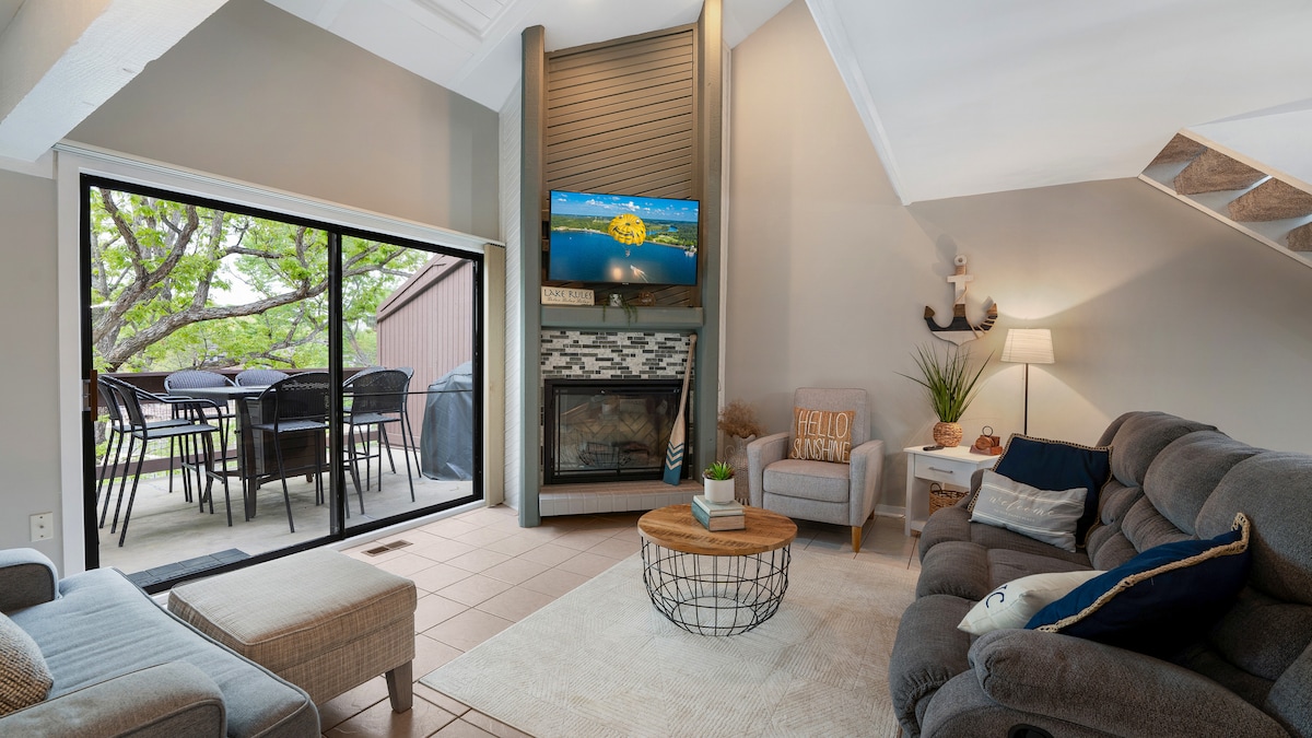 A cozy living area is highlighted by a large window offering views of the outdoors. A television is mounted above a modern fireplace, with a comfortable sofa and chair arranged around a round wooden coffee table. Natural light enhances the space.