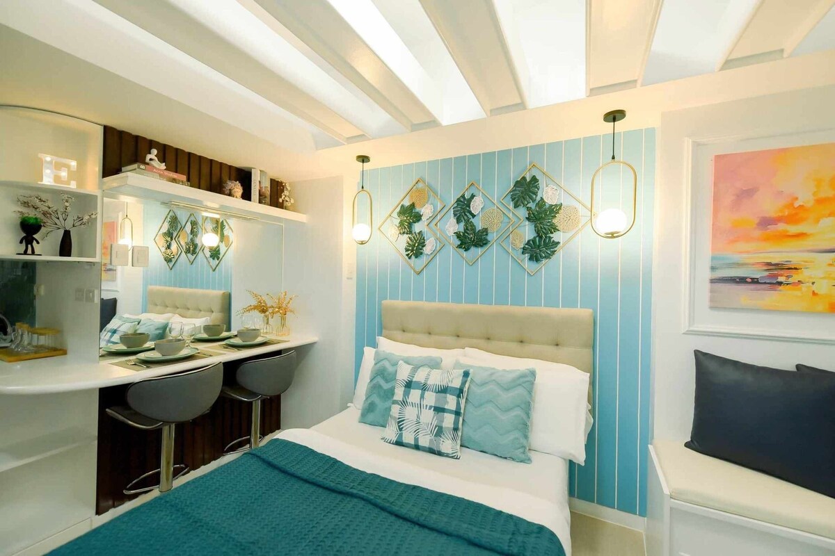 The bedroom area features a cozy bed with a tufted headboard, complemented by decorative pillows. A wall with blue stripes and botanical wall art enhances the ambiance. A compact dining space with bar stools is visible, along with natural light streaming in through the skylight.