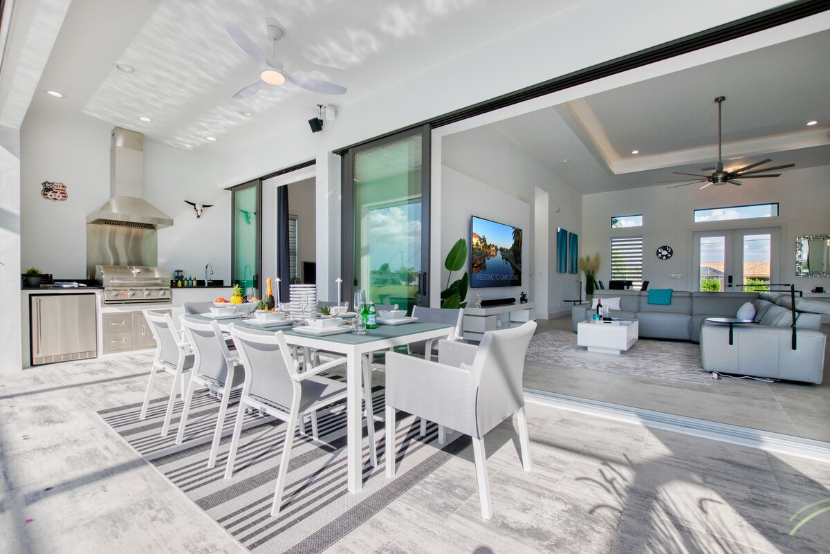 An outdoor dining area is featured under a covered lanai, equipped with a modern table and several white chairs. Clear glass doors provide a view into the spacious living area, which is brightened by natural light and includes a large sofa and a flat-screen TV.