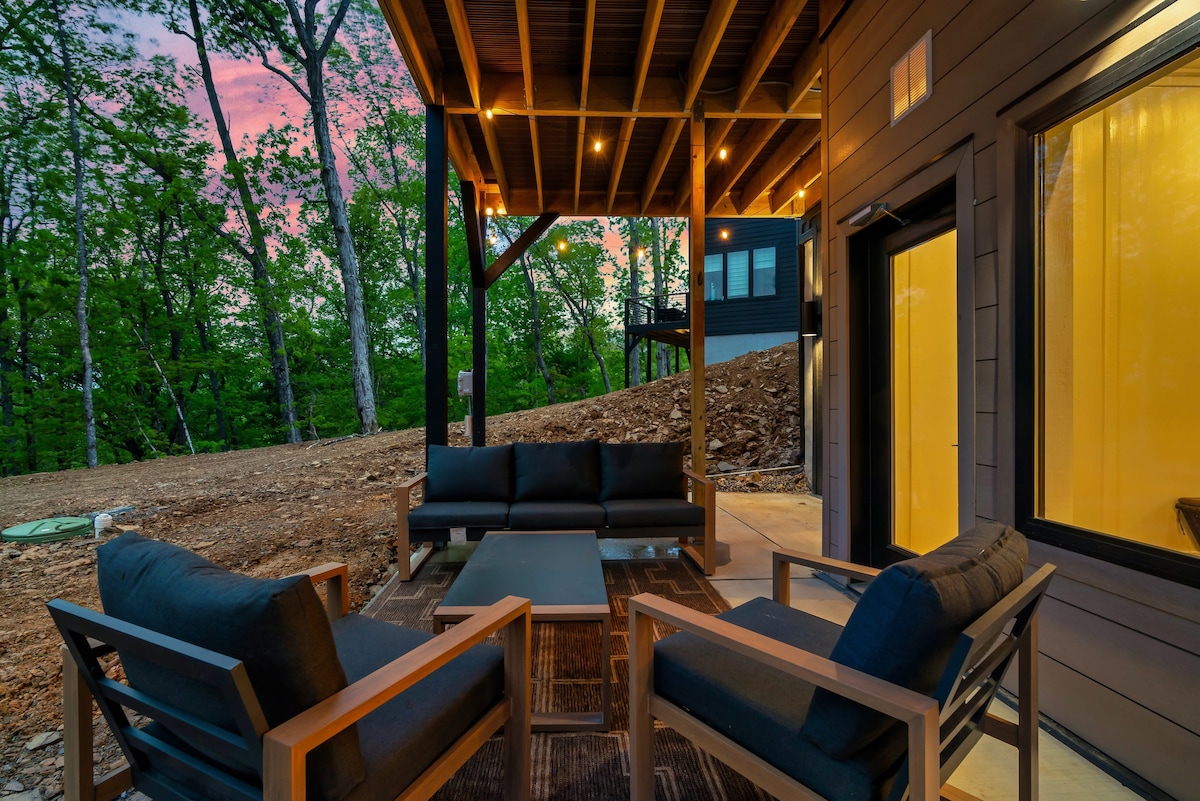 An outdoor seating area features comfortable couches and chairs arranged around a central coffee table, under soft lighting strung along the overhead beams. The natural surroundings with greenery are visible in the background, offering a serene atmosphere for relaxation.