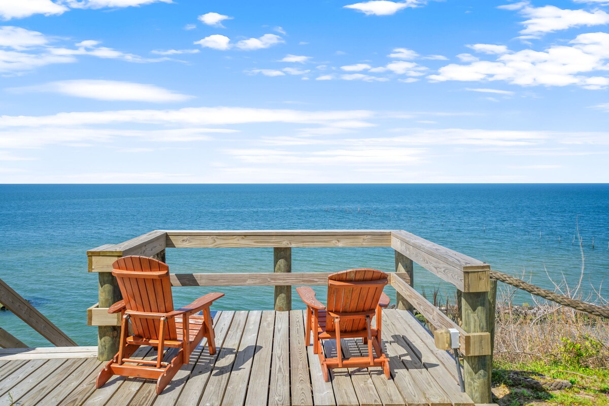 A wooden deck is shown overlooking the expansive Chesapeake Bay. Two rustic wooden chairs are positioned to face the water, providing an inviting spot for relaxation. The horizon is marked by a clear blue sky with scattered clouds, ensuring a serene coastal atmosphere.
