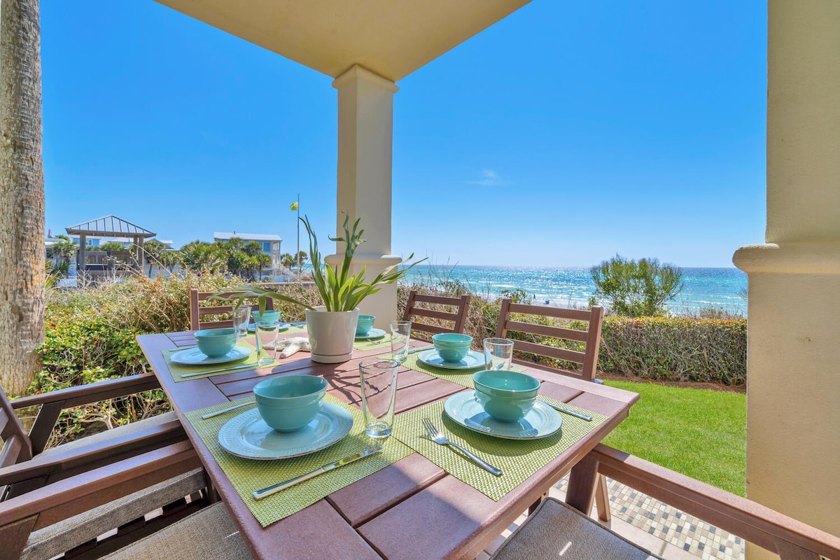 An outdoor dining table is set for a meal, featuring turquoise bowls and glassware. The lush green patio overlooks the Gulf of Mexico, with unobstructed views of the sparkling water and gentle waves. Sunlight brightens the scene, enhancing the beachfront ambiance.