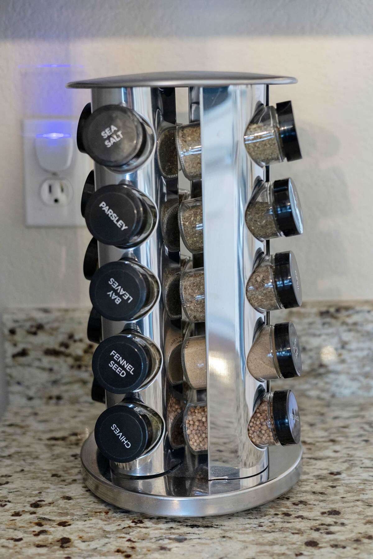 A rotating spice rack is displayed on a granite countertop, featuring sleek, labeled containers. Each container holds a different spice, with clear visibility of the contents. The modern design complements the kitchen's updated features.