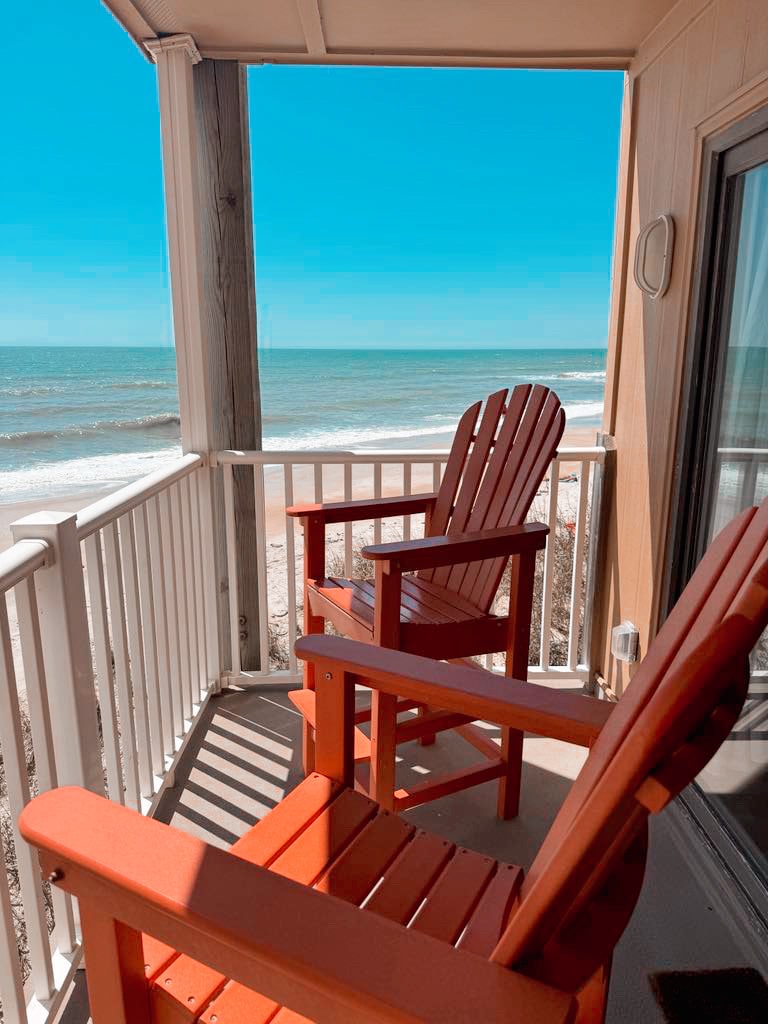 Two red Adirondack chairs are positioned on a covered patio overlooking the ocean. The clear blue sky complements the tranquil waves visible in the distance, creating a serene coastal scene.