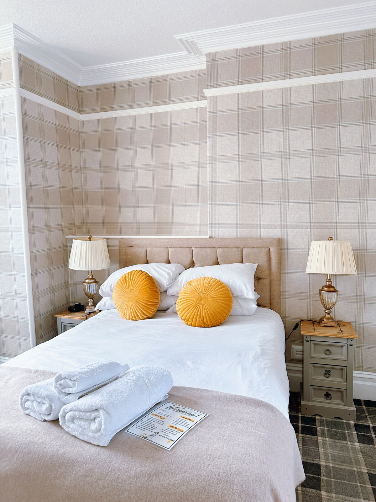 A cozy bedroom features a spacious bed dressed in soft linens, accented by two round, textured throw pillows. Two bedside tables with lamps flank the bed. A neutral plaid wallpaper adds a warm touch, and a folded towel set is neatly arranged nearby.