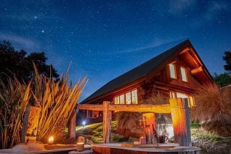 A rustic log chalet is illuminated under a starry sky, highlighting its wooden structure. Two outdoor baths are positioned in the foreground, surrounded by native plants. Soft lighting from candles adds warmth to the inviting scene while the natural surroundings create a serene retreat atmosphere.