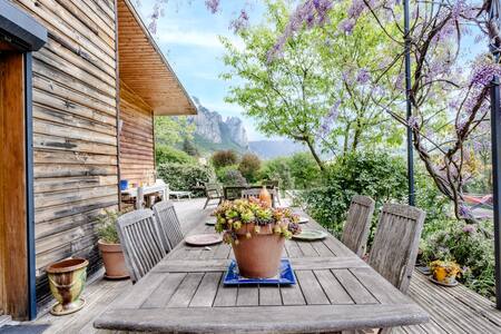 An outdoor dining area is featured, with a long wooden table surrounded by several wooden chairs. A potted plant sits at the center, and the scene is framed by blooming wisteria and lush greenery. Scenic hills can be seen in the background.