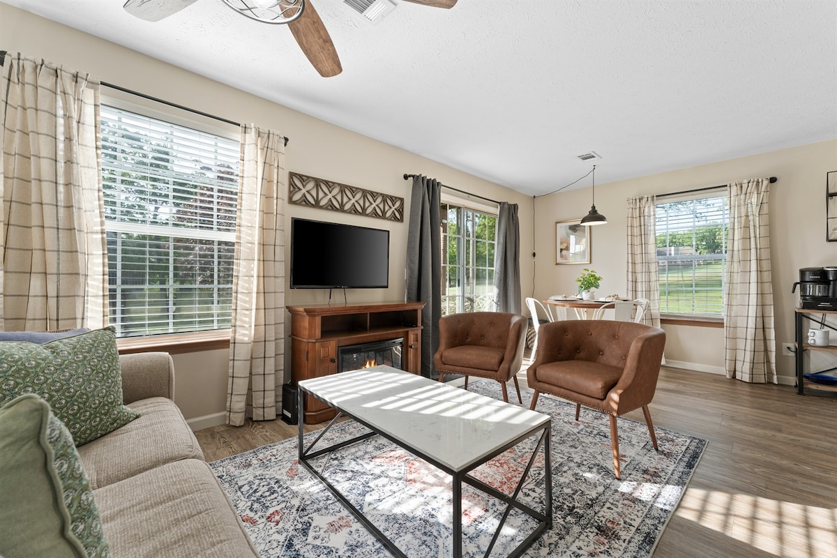 The living area features two brown chairs and a couch, arranged around a coffee table. A flat-screen television is mounted on the wall. Natural light filters through the windows, highlighting the cute decor and spacious layout, with a dining area visible in the background.