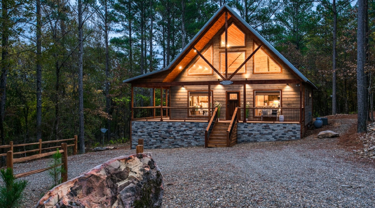 The cabin’s exterior is framed by tall trees, showcasing a warm wooden structure with a steeply pitched roof. A broad wooden deck extends from the front, featuring stairs leading to the entrance. Soft lighting highlights the cabin's inviting atmosphere.