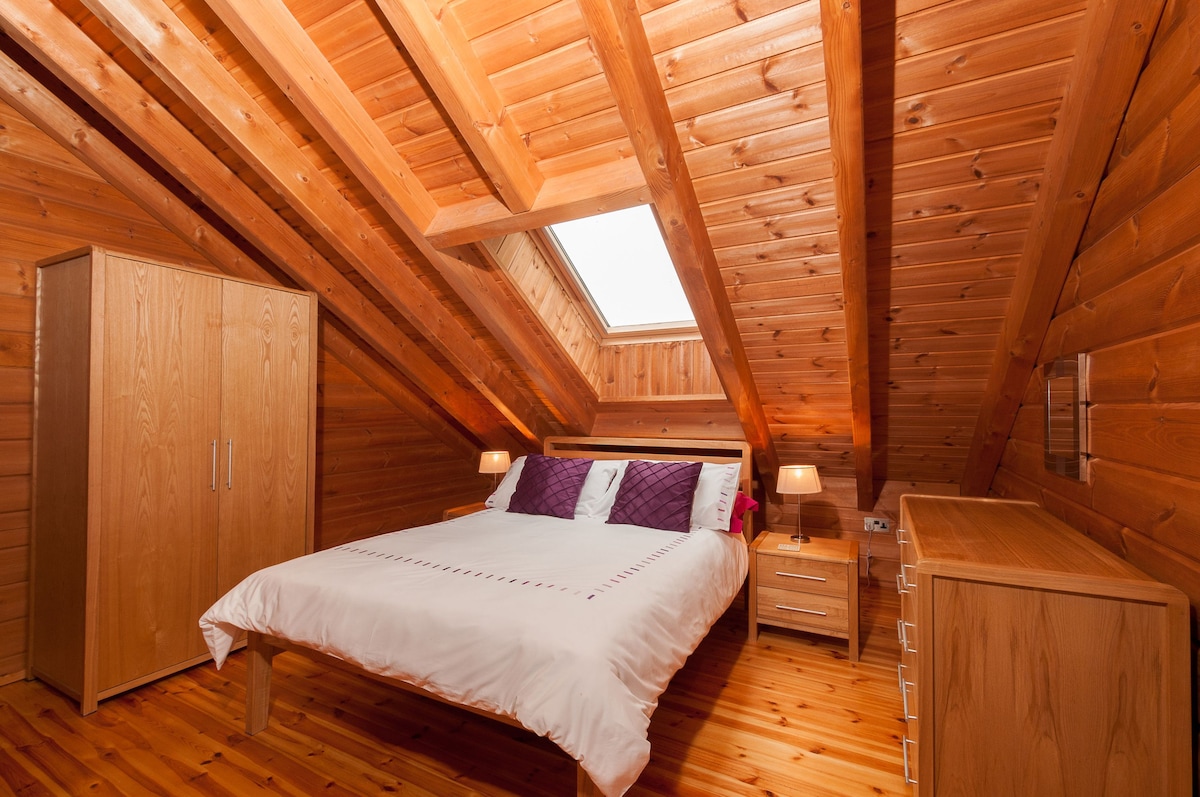 A serene bedroom is showcased with a sloped wooden ceiling and warm, natural lighting. The spacious bed is dressed in crisp white linens, featuring purple cushions. Flanking the bed are two bedside lamps, along with a wooden wardrobe and dresser, all harmonizing with the wood-paneled walls.