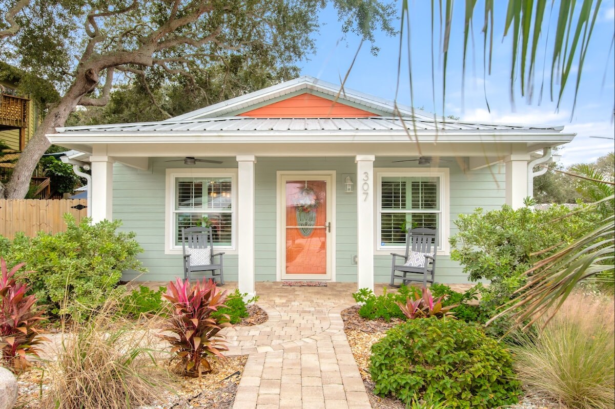 The exterior of a coastal-style home features a welcoming front path lined with tropical plants and decorative stones. A cozy porch with two rocking chairs invites relaxation, while the light green and coral color scheme complements the lush surroundings.