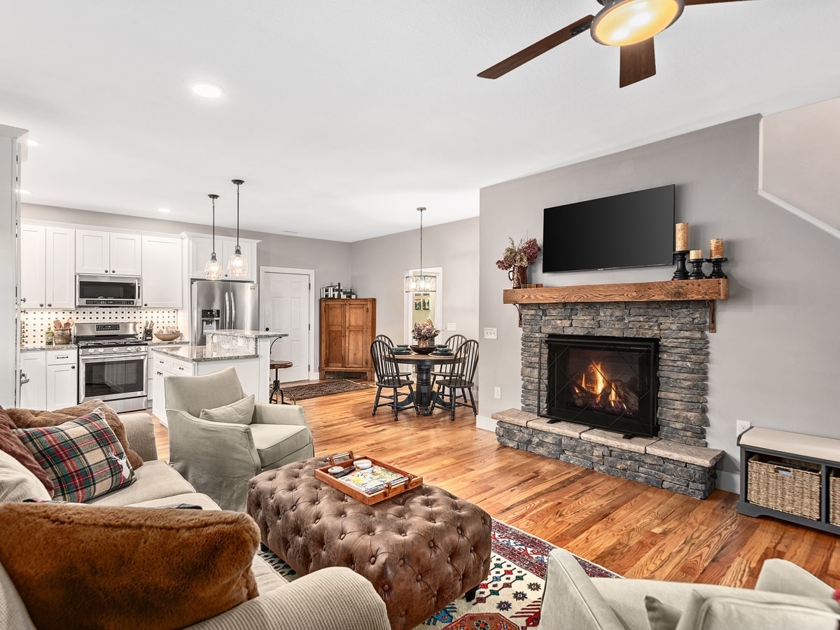A living area features a comfortable seating arrangement with plush armchairs and a large ottoman. A stone fireplace is situated below a wall-mounted television. A wooden dining table with chairs is visible in the background, complemented by warm wood tones and soft lighting.