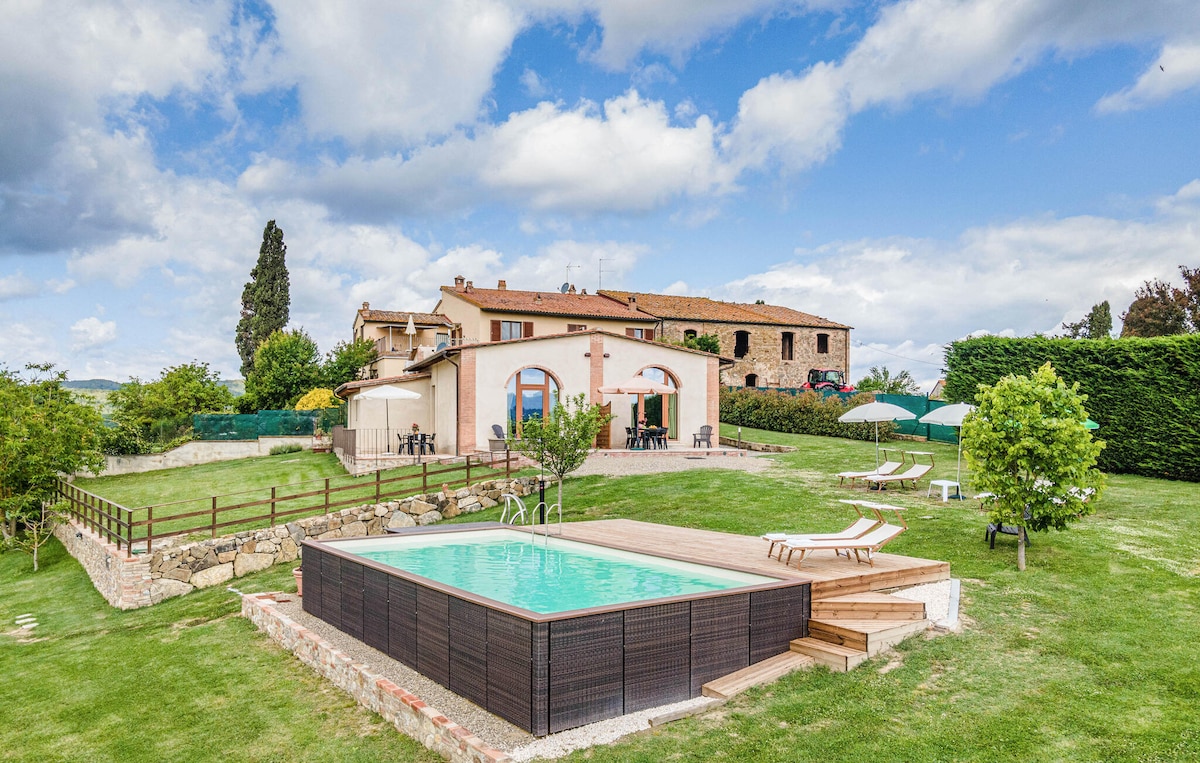 A shared outdoor pool sits in a spacious grassy area, surrounded by trees and lounging chairs. A two-story apartment building is visible in the background, featuring a mix of arch and rectangular windows. Umbrellas provide shade beside the pool, enhancing the inviting outdoor space.