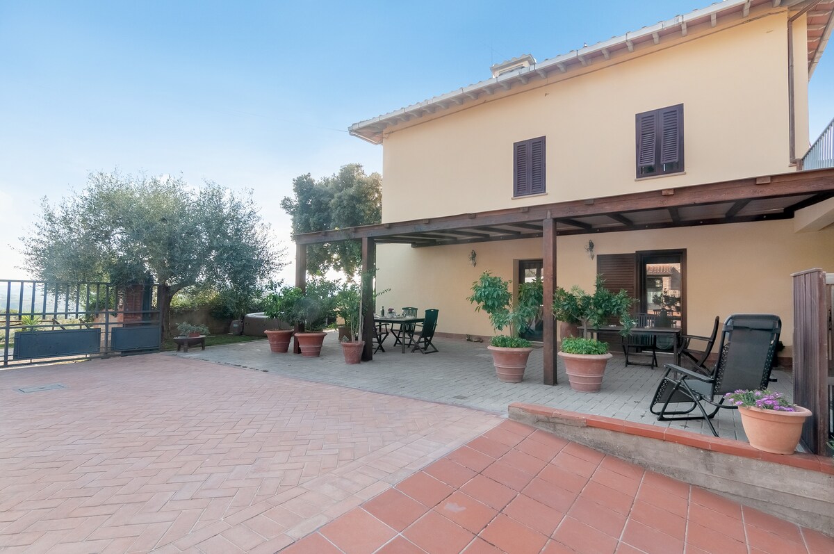 An outdoor terrace is featured, with potted plants and shaded seating areas. The structure displays a light-colored facade with dark shutters, and a spacious layout invites relaxation in a scenic hillside setting.