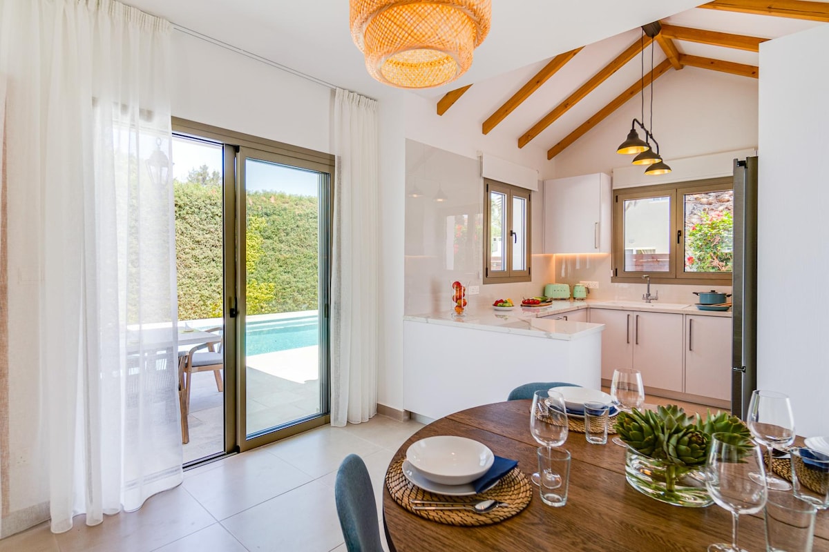 An open-plan kitchen and dining area features a round wooden table set for four, complemented by soft lighting from a woven pendant. Large sliding glass doors provide a view of the pool and backyard, while natural light floods the space through soft, sheer curtains.