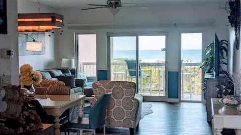 A spacious living area is highlighted by large glass doors that open to reveal ocean views. Comfortable seating is arranged around a coffee table, with a ceiling fan overhead. Light wood flooring complements the bright decor, creating a welcoming atmosphere.