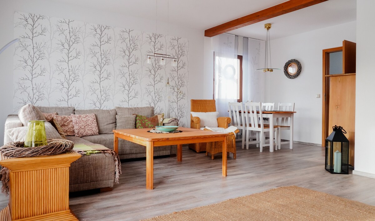 A spacious living area features a comfortable sofa with textured cushions and a central coffee table. A dining table is visible, surrounded by white chairs. Natural light illuminates the space, highlighting the light wood flooring and decorative wall patterns.