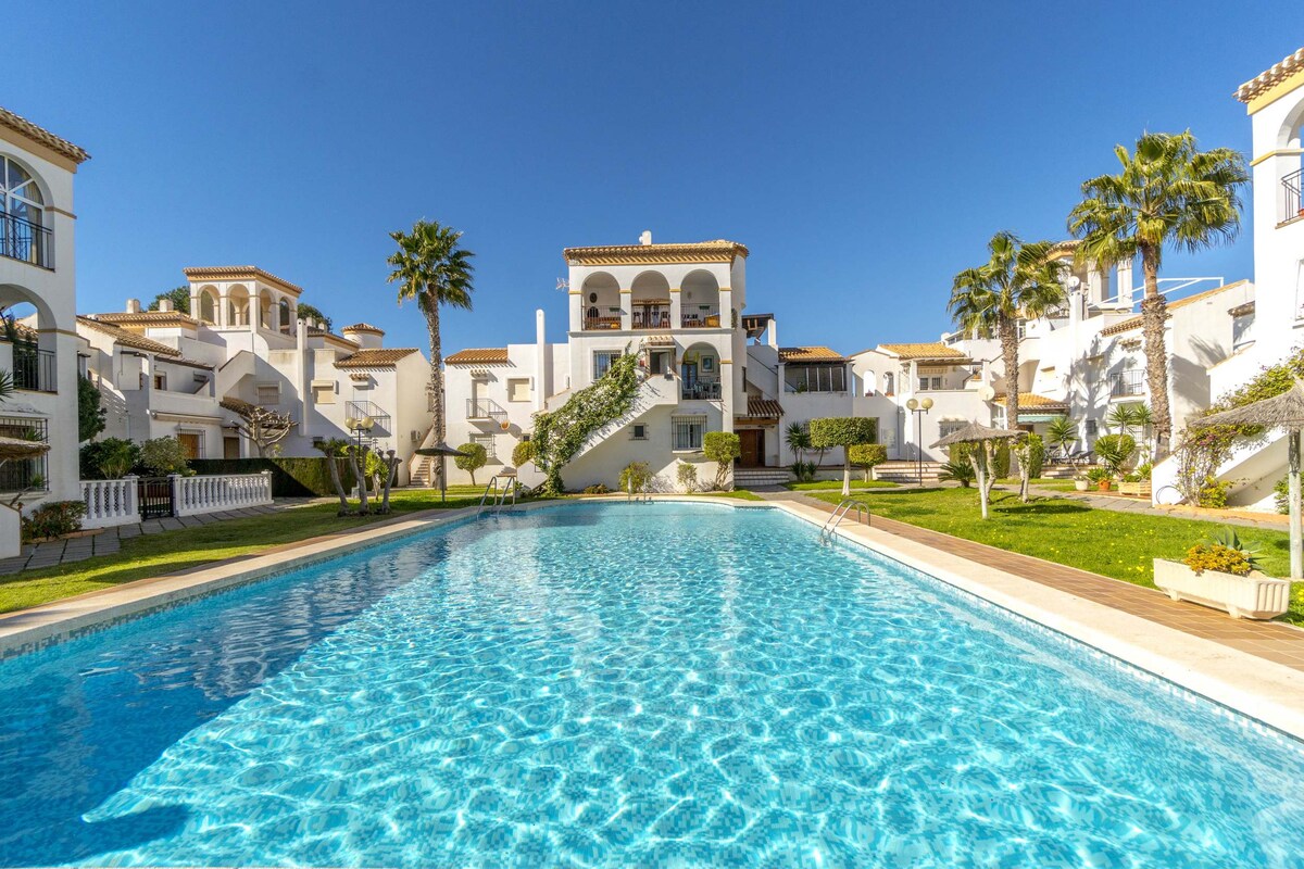 The image showcases a large swimming pool surrounded by well-manicured gardens and palm trees. The bright blue water invites relaxation, while the Mediterranean-style buildings rise in the background, creating a serene and spacious communal area.