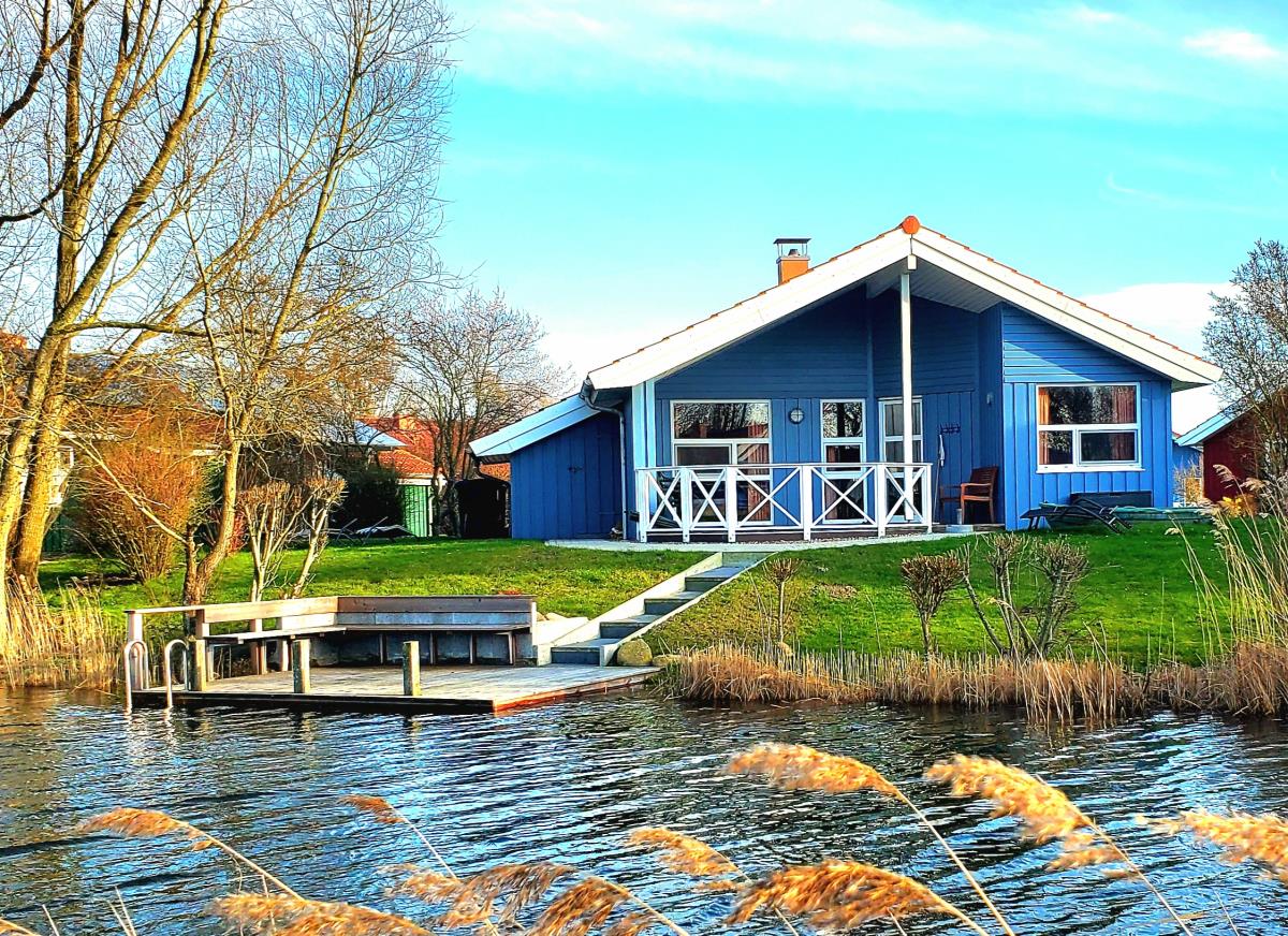 A blue wooden house is situated near the water's edge, with a private dock extending into the lake. Lush grass and slender trees frame the property, creating a serene environment. Wide windows reflect natural light, and a small staircase leads from the house to the dock.