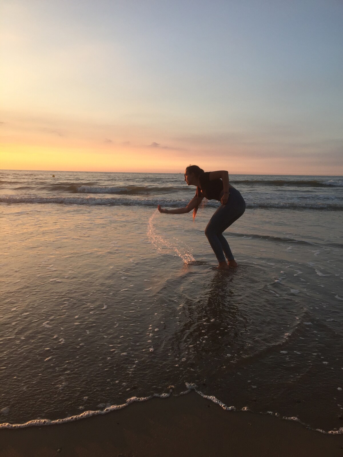 The image captures a person engaging playfully with the gentle waves at the beach during sunset. The sky is painted with soft hues of orange and purple, while the reflection of the water's surface adds a serene ambiance to the coastal scenery.