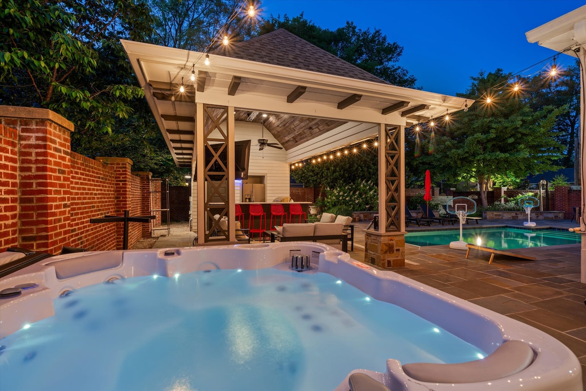 A serene outdoor retreat features a hot tub with glowing water, surrounded by stone flooring. A shaded area is adorned with string lights and offers comfortable seating. The private pool reflects the evening sky, enhancing the relaxed ambiance of the space.