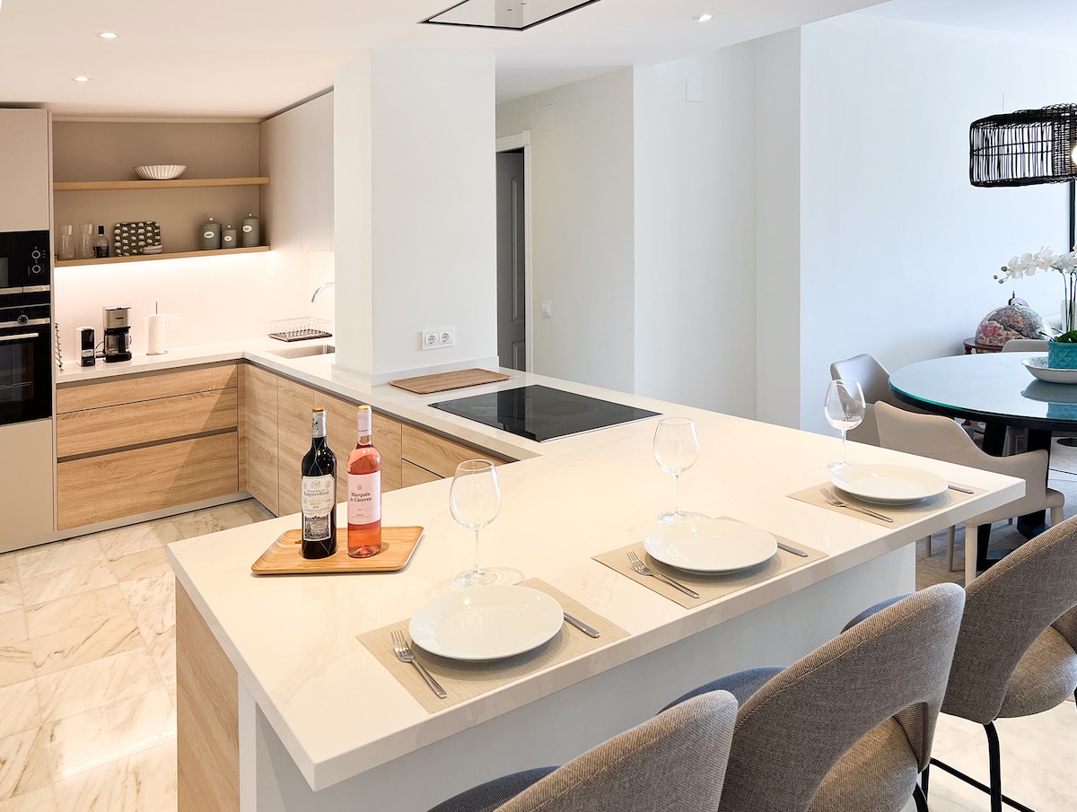 A modern open-plan kitchen features light wood cabinetry and white countertops. An island counter is set for three, displaying plates and wine glasses. A dining area is visible in the background, showcasing a round table with additional seating. Soft lighting enhances the warm ambiance.