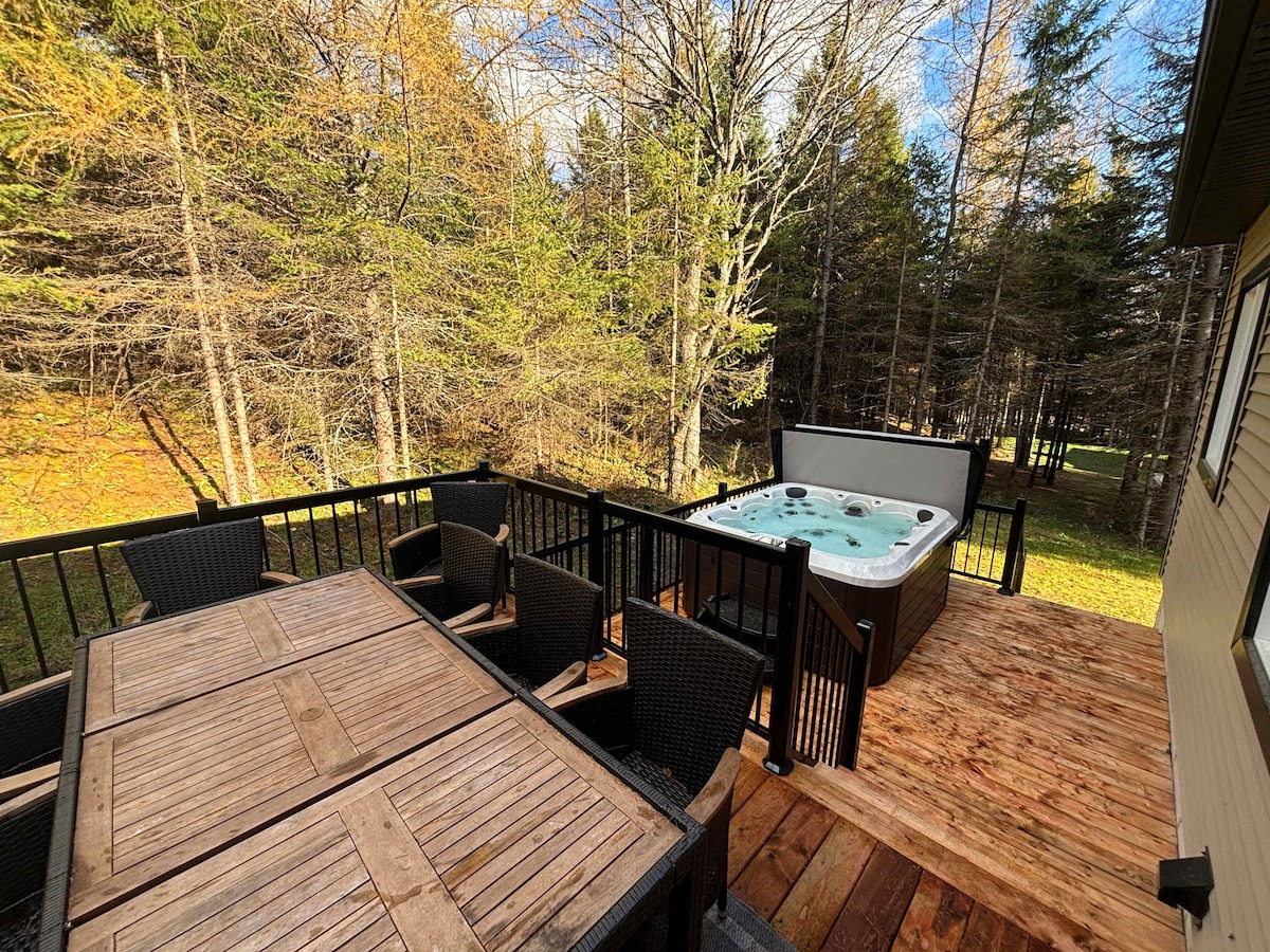 An inviting outdoor terrace is highlighted by a large wooden dining table surrounded by six black chairs. A hot tub is positioned nearby, nestled within a natural setting of trees, creating a serene space for relaxation.