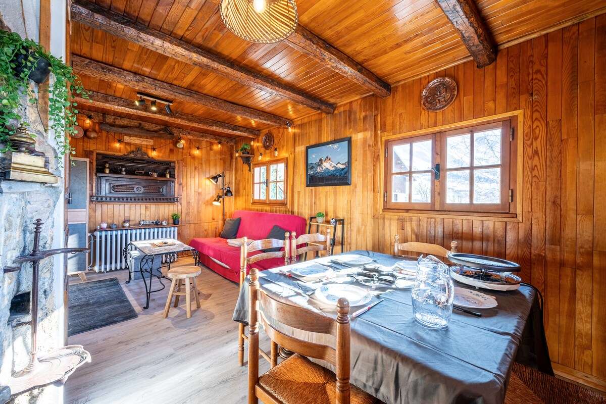 A warm and inviting living space is highlighted by wooden paneling and exposed beams. A spacious sofa faces a functional fireplace, while a dining table is set for four. Natural light pours in through windows, creating a bright and cozy atmosphere.
