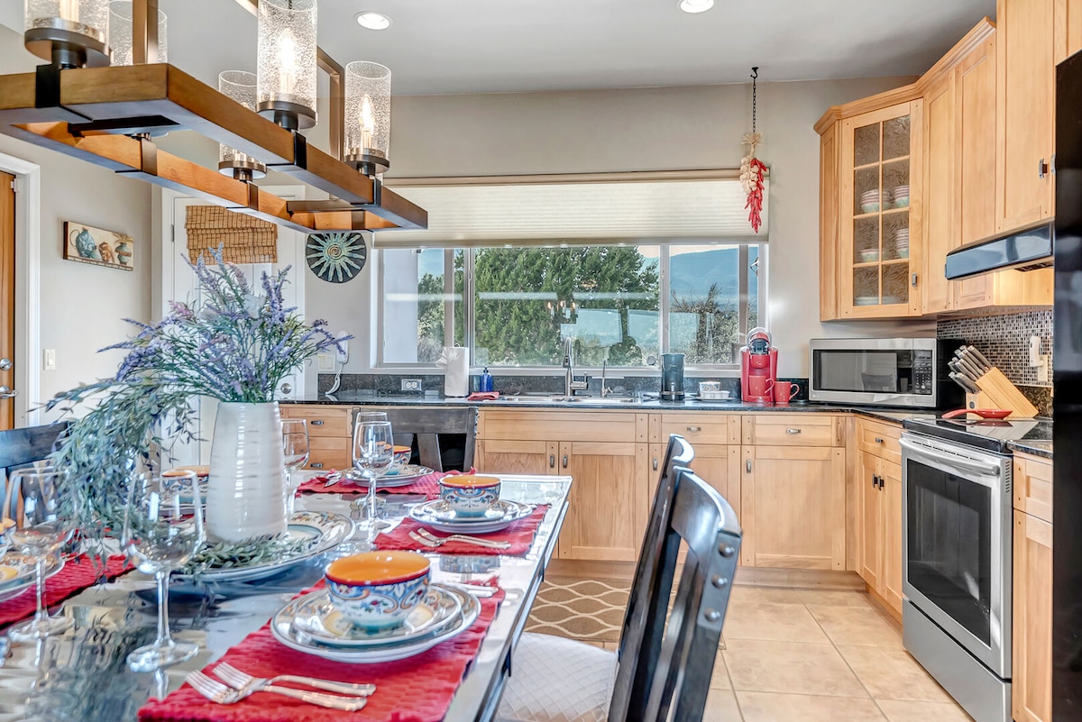 A well-equipped kitchen features natural wood cabinetry and a spacious dining area with a large table set for six. Bright light fills the room through wide windows, showcasing views of the surrounding landscape. Modern appliances and elegant tableware are arranged for a welcoming dining experience.