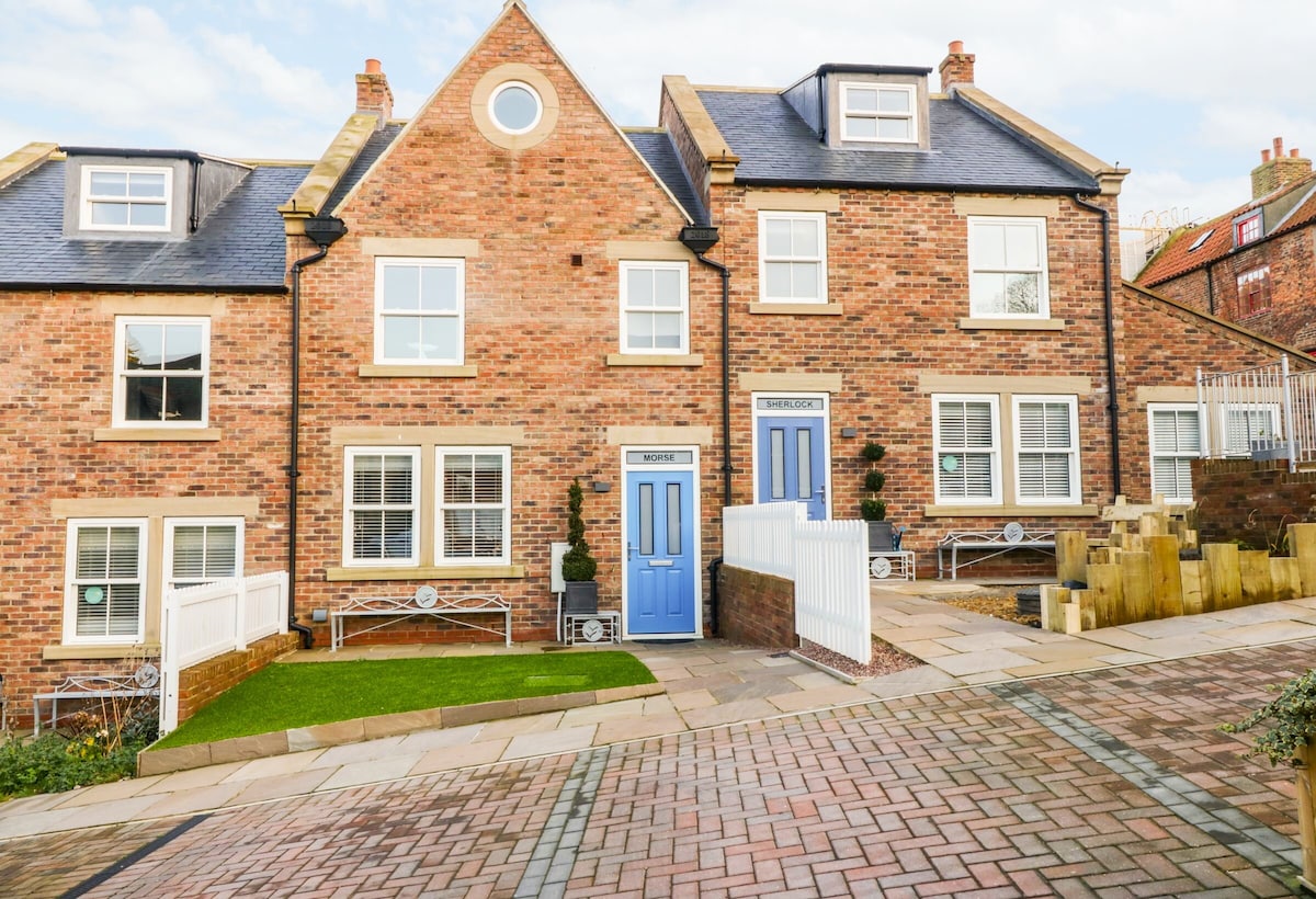 The exterior view of Morse Cottage reveals a charming brick façade with a pitched roof. Two blue doors provide a welcoming entrance, flanked by large windows. The landscaped area includes green grass and stone pathways, adding to the inviting atmosphere of the property.