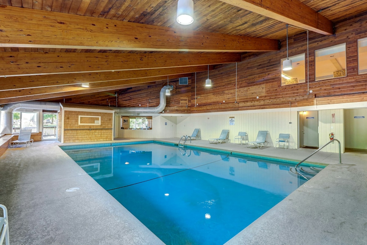 An indoor swimming pool is surrounded by wooden beams and a light-colored ceiling, creating a spacious atmosphere. Several lounge chairs are placed along the side, and large windows provide natural light and views of the outdoors.