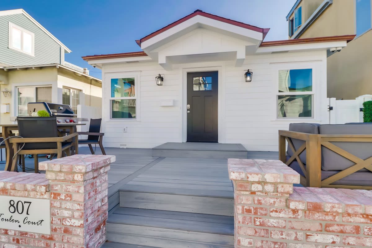 The charming exterior of the cottage features a welcoming front porch, complete with comfortable seating and a barbecue grill. A pathway leads to the stylish front door, framed by several large windows that allow for natural light. The contrast of white siding and red roof adds to the coastal appeal.