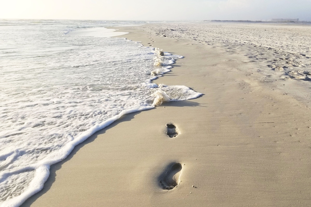 A serene beach scene is depicted, featuring soft sand with gentle waves lapping at the shore. Footprints are visible leading along the water's edge, where the foamy surf meets the pale sand, inviting a sense of calm and tranquility.