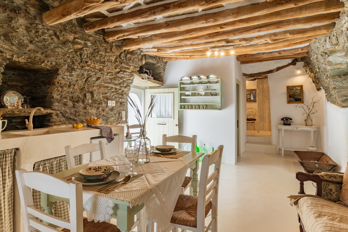 A charming dining area features a light-colored wooden table set with dishes and glassware. The rustic stone walls are complemented by exposed wooden beams overhead. A small kitchen space is visible, adding to the homely atmosphere of the room.
