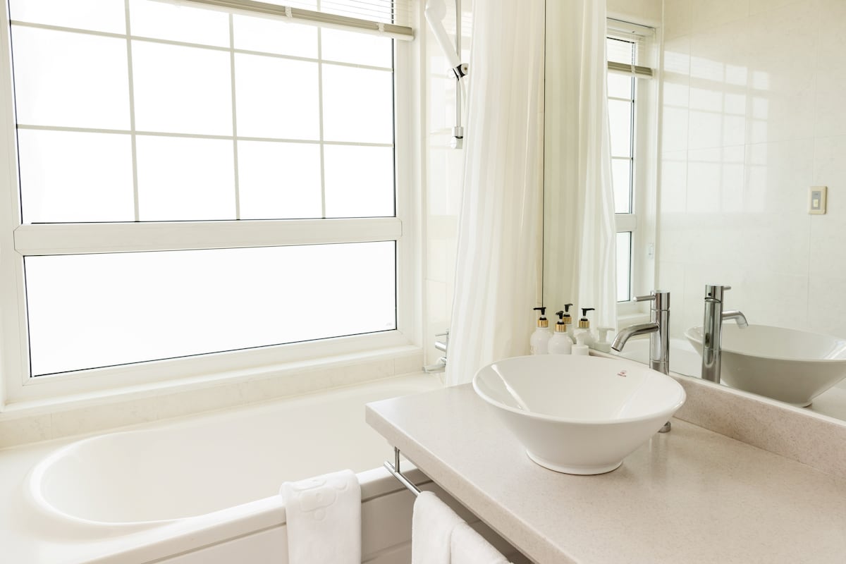 A bright bathroom space is equipped with a large bathtub and a modern sink. Natural light enters through a spacious window with grid patterns, complemented by soft white curtains. A clean, light-colored countertop is paired with neatly arranged toiletries.