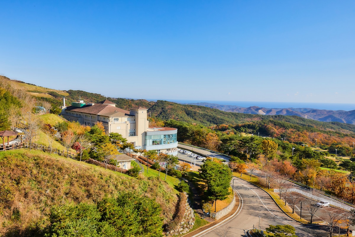 The image features a resort-style building situated on a hillside, surrounded by lush greenery and colorful autumn foliage. A winding road leads up to the entrance, with expansive views of distant mountains under a clear blue sky.