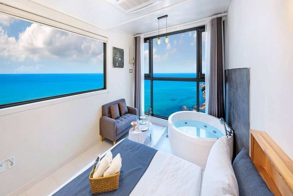 A spacious room features a comfortable bed adorned with soft linens, a cozy seating area with a gray sofa, and a stylish round bathtub. Large windows provide an expansive ocean view, flooding the space with natural light.