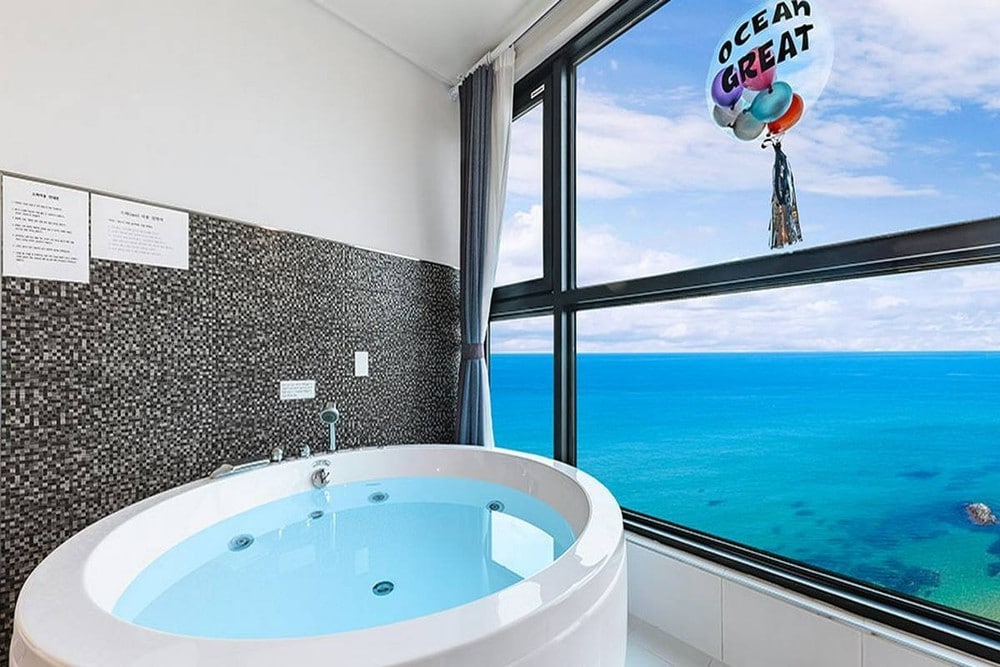 A round, private spa tub is positioned next to large windows that offer expansive views of the ocean. The soothing blue water of the tub is complemented by the mosaic tile wall, while the distant horizon meets the sky.