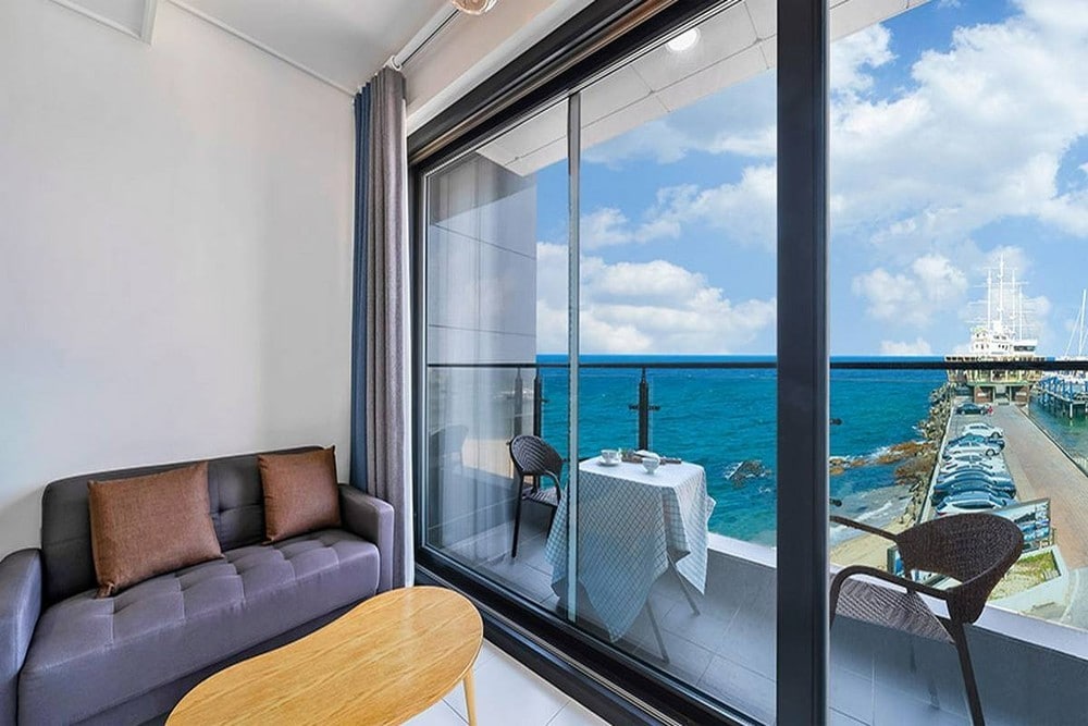 A spacious living area features a comfortable gray sofa and a wooden coffee table. Large glass doors open to a balcony with a stunning ocean view, showcasing boats in the water. A small table with two wicker chairs is set for outdoor relaxation.