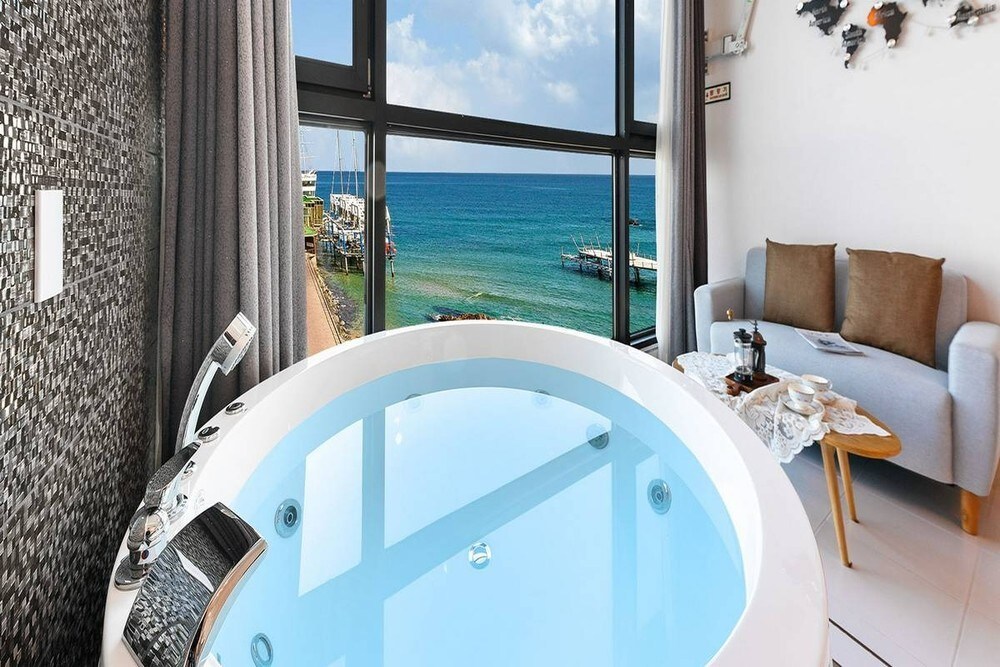 A private spa bath is positioned near a large window, offering an unobstructed view of the ocean. Soft gray curtains frame the scene, while a comfortable seating area with a light-colored sofa and a small table is visible in the background.