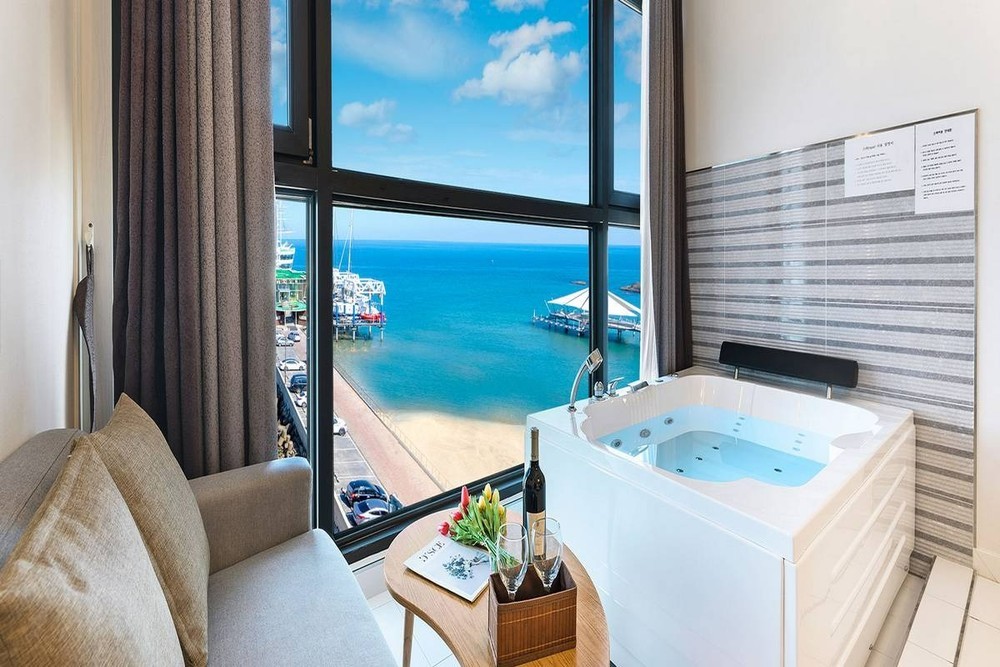A spacious room features a large window revealing an ocean view. A private spa bath is positioned next to the window, complemented by a small table set with a bottle of wine and a floral arrangement. A gray sofa is situated nearby.