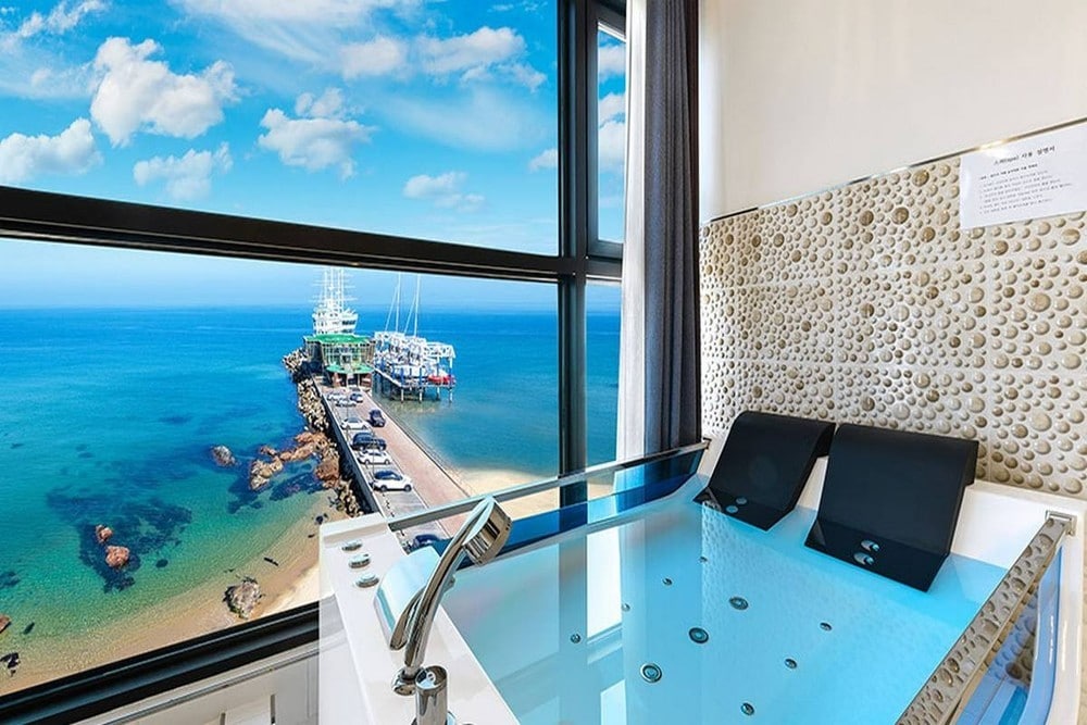 A private spa is situated beside large windows that offer a panoramic view of the ocean and pier. The spa is equipped with sleek black seating, and the soothing water features bubbles and jets, inviting relaxation against the backdrop of the tranquil seascape.