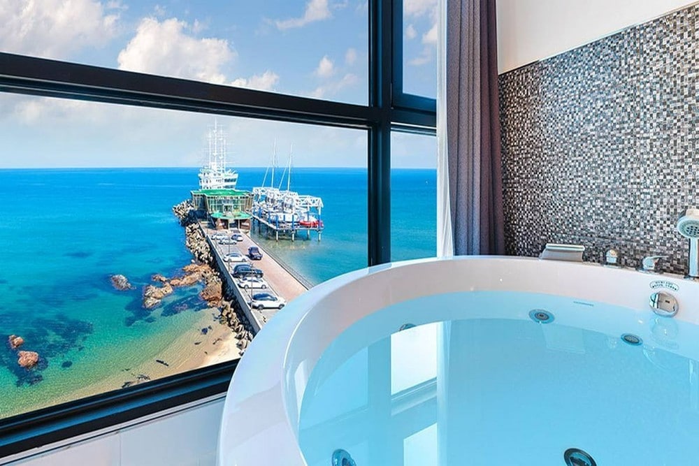 A private spa tub is positioned next to large windows, offering a view of the ocean and pier. The water in the tub reflects the calm blue hue of the sea, while mosaic tiles embellish the wall, enhancing the serene ambiance.