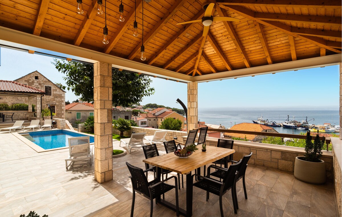 A spacious covered terrace is highlighted by a large wooden dining table surrounded by black chairs. A pool glistens nearby, with views of the sea and charming coastal houses in the background. Ceiling fans are visible, providing a cool breeze.