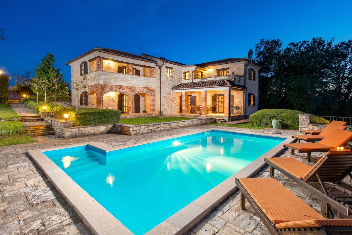 An outdoor view of a rustic stone house is illuminated against the evening sky. A turquoise swimming pool rests in the foreground, surrounded by landscaped greenery. Sun loungers with orange cushions are positioned beside the pool, offering a relaxing space in a tranquil setting.